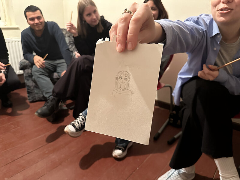 Eine Person hält ein Blatt Papier mit einer gezeichneten Karikatur vor. Im Hintergrund sitzen weitere Personen, die ebenfalls zeichnen. Die gezeichnete Figur hat große Augen und wirkt leicht übertrieben. Die Szene scheint in einem kreativen Workshop oder einer Zeichnungsession stattzufinden.