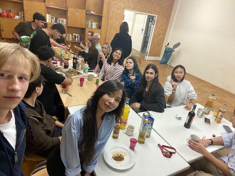 Gruppe von Menschen sitzt in einem Raum an langen Tischen, isst gemeinsam und unterhält sich. Auf den Tischen stehen Gläser, Flaschen, Teller mit Essen und Snacks. Gemütliche Atmosphäre mit vielen Personen im Hintergrund.