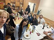 Gruppe von Menschen sitzt in einem Raum an langen Tischen, isst gemeinsam und unterhält sich. Auf den Tischen stehen Gläser, Flaschen, Teller mit Essen und Snacks. Gemütliche Atmosphäre mit vielen Personen im Hintergrund.