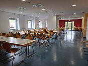 Ein leerer Speisesaal mit mehreren Tischen und Stühlen. Die Tische sind in Reihen angeordnet, und die Stühle sind an den Tischen platziert. Der Raum hat große Fenster an einer Wand und eine Tür am anderen Ende. Die Wände sind weiß, und der Boden ist dunkelgrau.
