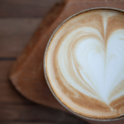 Ein Tassenrand mit einem kunstvoll gezeichneten Herz aus Milchschaum. Die Tasse steht auf einer Holzunterlage. Der Milchschaum ist hell und bildet ein deutliches Herzmuster.