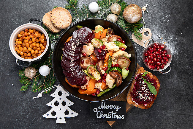 Weihnachtliches Festmahl mit gebratenem Gemüse wie Pastinaken, Karotten und Pilzen in einer Pfanne, dazu geröstete Kichererbsen, Cranberries, geräucherter Lachs auf Toast und ein Tannenbaum-Motiv mit der Aufschrift „Merry Christmas“.