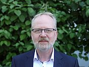 Ein älterer Mann mit grauen Haaren, Brille und grauem Bart trägt ein blaues Sakko und ein weißes Hemd. Er steht vor einem grünen, belaubten Hintergrund.