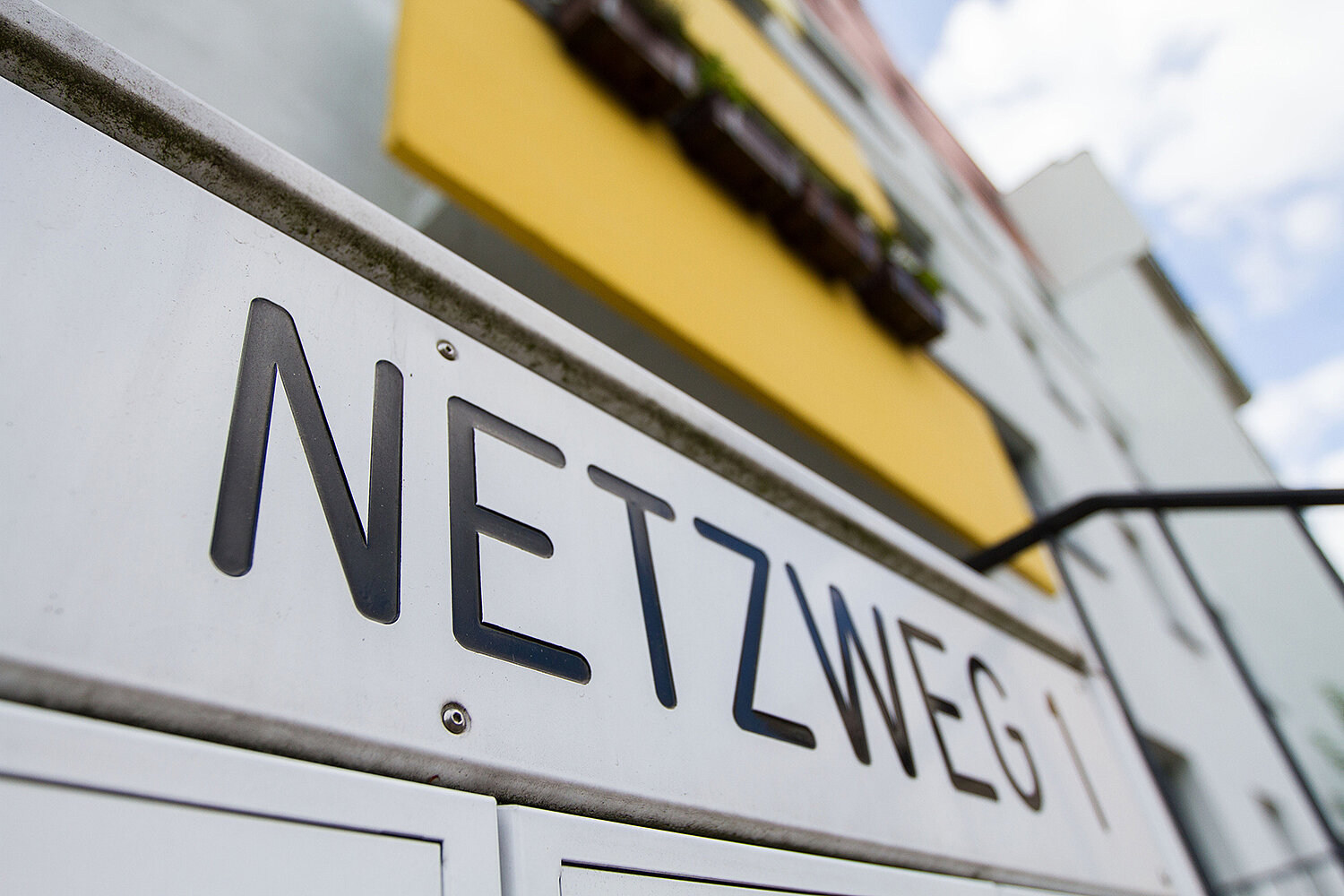 "Ein Straßenschild mit der Aufschrift 'NETZWEG' in Großbuchstaben. Das Schild ist weiß mit schwarzer Schrift und befindet sich an einem Gebäude mit gelbem Fensterrahmen."