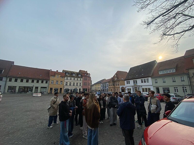 Eine Gruppe von Menschen steht auf einem Platz in einer historischen Stadt. Die Gebäude im Hintergrund haben traditionelle europäische Architektur mit roten Dächern und hellen Fassaden. Die Atmosphäre wirkt lebhaft und gesellig.