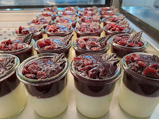 Drei Reihen mit kleinen Gläsern, gefüllt mit zweilagigem Dessert: unten cremige weiße Schicht, oben dunkle Schokoladencreme, garniert mit frischen Himbeeren und Schokoraspeln, teilweise mit Schokoladenblättern verziert.