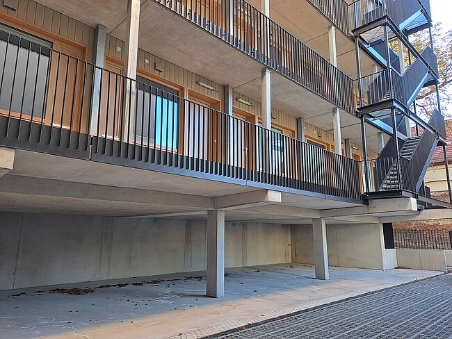 Moderner Mehrfamilienwohnhausbau mit mehreren Geschossen, die durch Geländer abgesicherte Balkone und große Fenster aufweisen. Im Vordergrund eine ebenerdige, offene Stellfläche mit Betonfundament und Treppe zum oberen Eingang.