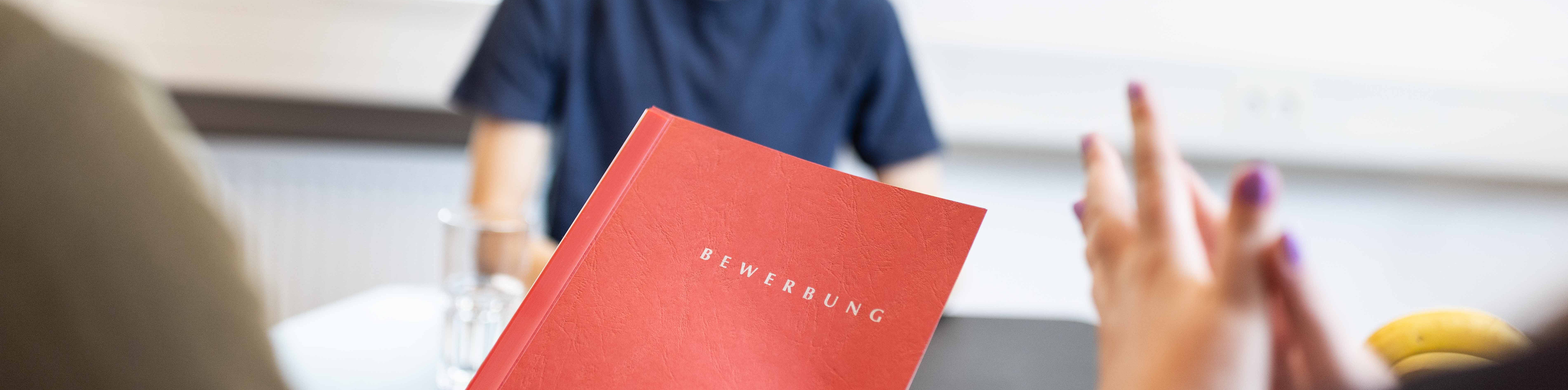 Eine Person hält einen roten Ordner mit der Aufschrift „Bewerbung“ in einem Vorstellungsgespräch hoch, während im Hintergrund weitere Teilnehmer an einem Tisch sitzen und diskutieren.