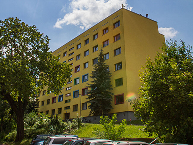 Ein sonniger Tag mit einem hellen, strahlenden Himmel. Im Vordergrund steht ein mehrstöckiges, gelbes Gebäude mit vielen Fenstern. Um das Gebäude herum sind Bäume und Sträucher zu sehen.