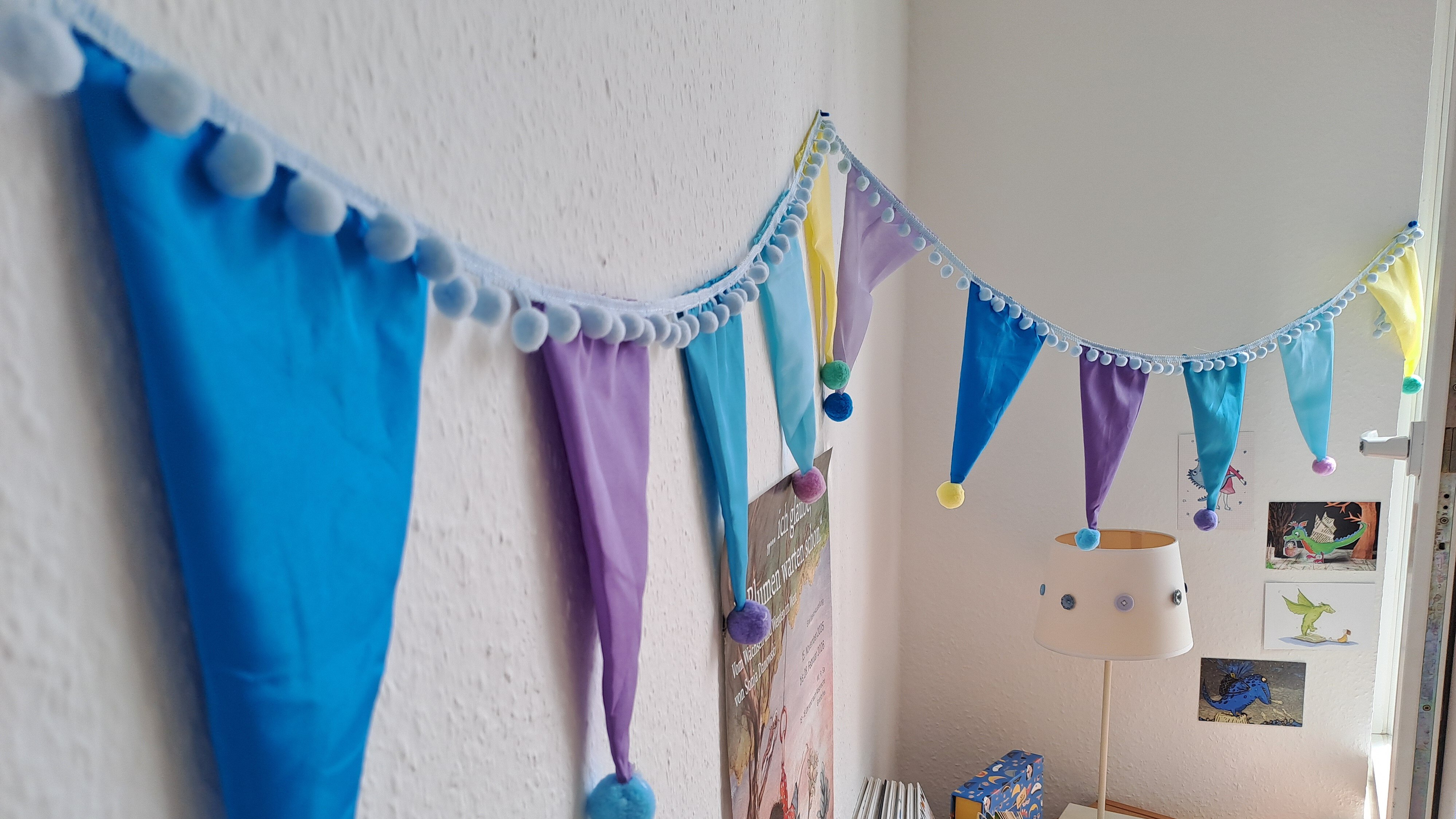 Bunte Girlande mit dreieckigen, pastellfarbenen Bändern in Blau, Violett und Pink an einer Wand aufgehängt, verziert mit kleinen Pompons und Glöckchen. Im Hintergrund ein Kinderzimmer mit Regal, Büchern und Spielzeug.