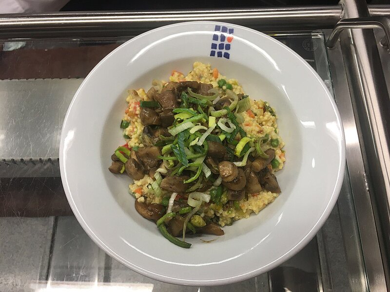 Ein weißer Teller mit einer Mahlzeit aus Quinoa, Pilzen und grünen Zwiebeln. Die Quinoa ist gelblich und die Pilze sind braun. Die grünen Zwiebeln sind fein geschnitten und über das Gericht verteilt.
