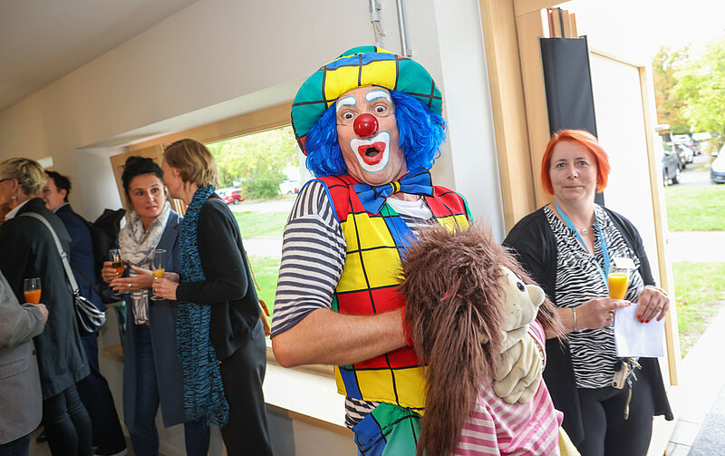 Ein Clown in bunter Kostüm steht in einem Raum und hält ein Kind an der Hand. Um sie herum stehen mehrere Erwachsene, die Getränke in den Händen halten und sich unterhalten. Die Atmosphäre wirkt fröhlich und gesellig.