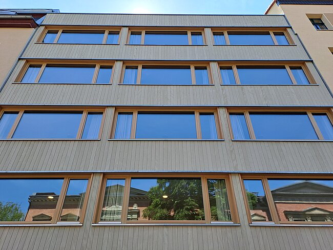 Moderne viergeschossige Fassade mit vertikalen Holzlamellen, großen Glasfenstern und Spiegelungen von Bäumen und Himmel.