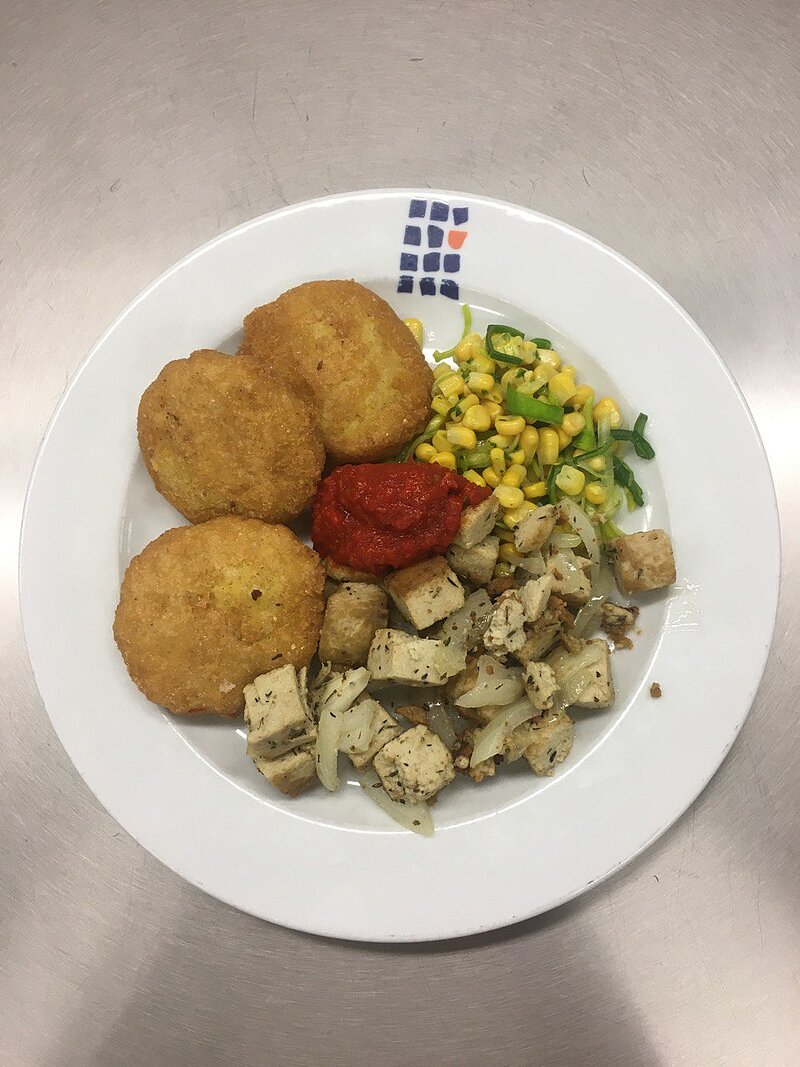 Ein Teller mit vier goldbraunen Frikadellen, einer Portion Mais mit grünen Paprikastücken, gebratenem Tofu und Kartoffelwürfeln. Dazu gibt es eine kleine Portion Ketchup.
