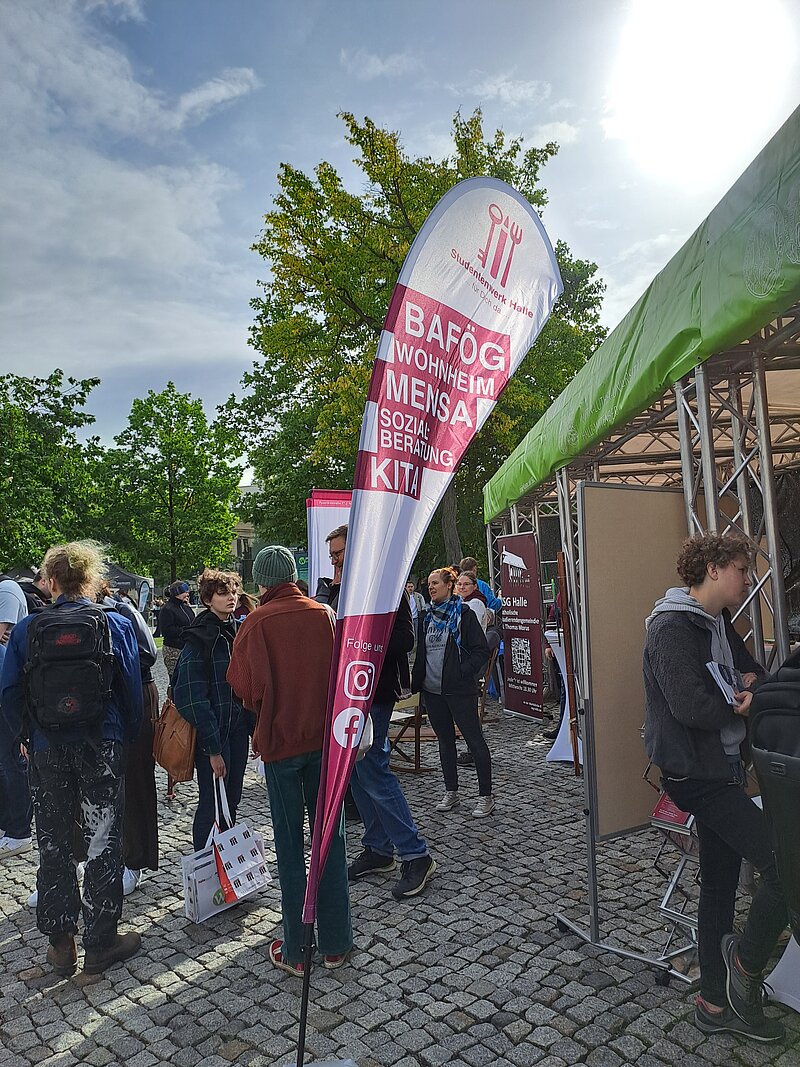 "Menschen stehen in einer Schlange vor einem Imbisswagen. Ein Schild mit der Aufschrift 'Bafög' und 'Mensa' ist sichtbar. Die Szene spielt im Freien, möglicherweise auf einem Campus oder bei einem Event. Die Atmosphäre wirkt lebhaft und informell."