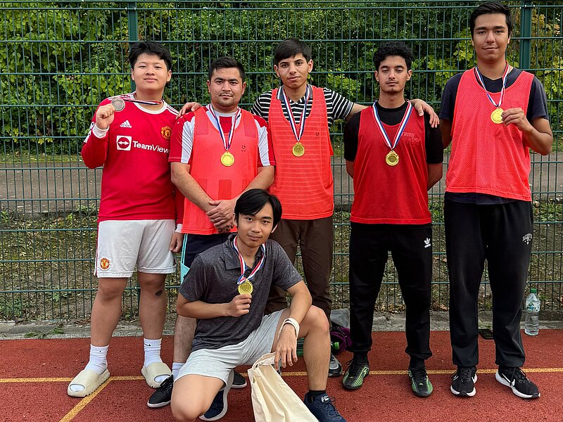 Fünf Personen in roten Trikots mit Medaillen um den Hals stehen auf einem Tennisplatz und posieren stolz. Eine Person kniet vor ihnen und hält eine Trophäe. Sie feiern gemeinsam einen Sieg.