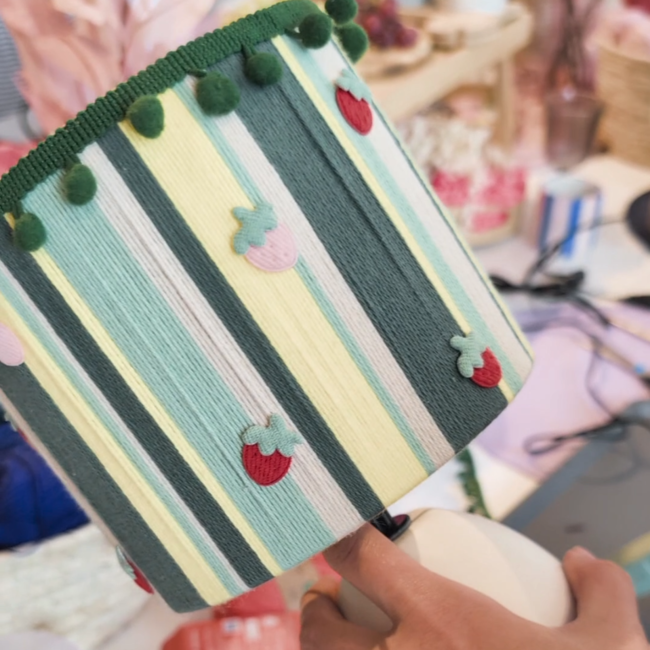 Lampe, Foto: Anna Möbius, bunt&BÄM Bunt gestreifter, handgefertigter Lampenschirm aus Stoff mit grünen Bordüre und aufgenähten kleinen Stofferdbeeren in Rot und Pink, gehalten von einer Hand.