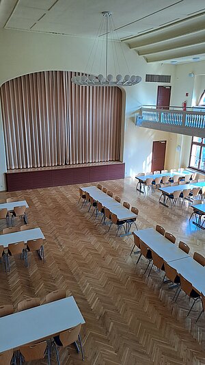 Ein leerer Speisesaal mit mehreren Tischen und Stühlen, die in Reihen angeordnet sind. An einer Wand befindet sich ein großer Vorhang, und im Hintergrund ist eine Galerie mit einem Geländer zu sehen. Die Decke hat eine moderne Beleuchtung. Der Boden ist mit Holzparkett ausgelegt.