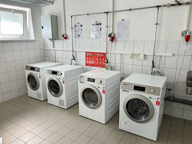 Vier nebeneinander aufgestellte Waschmaschinen und Trockner in einem Technikraum, alle mit Bedienfeld und Sicherheitshinweisen.