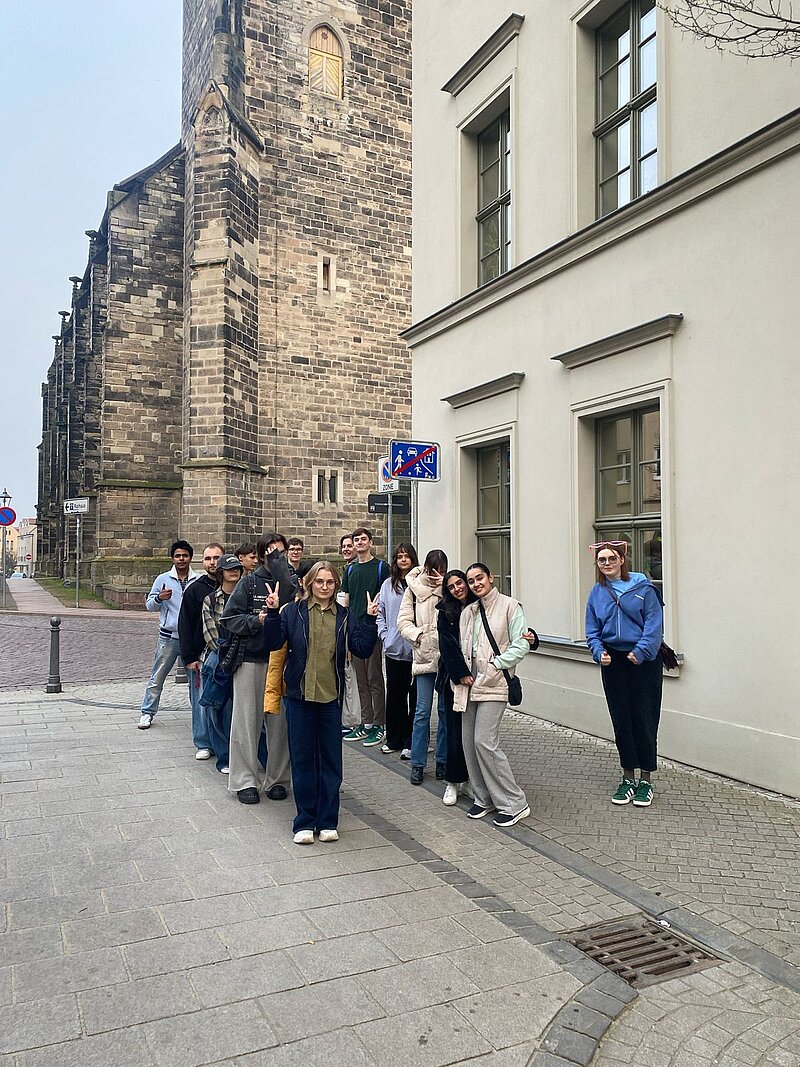 Eine Gruppe von Menschen steht vor einem alten Gebäude mit Steinmauer und einem modernen Gebäude mit Fenstern. Sie scheinen auf einer Stadtführung oder einem Ausflug zu sein. Die Straße ist gepflastert und es gibt ein blaues Straßenschild in der Nähe.