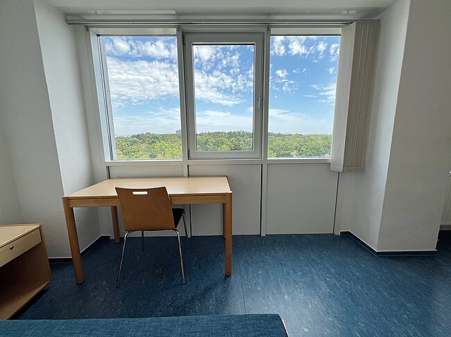 Ein helles, schlichtes Zimmer mit großem Fenster, das Blick auf blauen Himmel mit Wolken und grünes Gelände bietet. Vor dem Fenster steht ein kleiner Holztisch mit einem Stuhl.