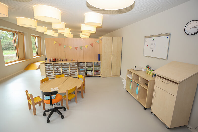 Ein heller, moderner Klassenraum mit einem runden Tisch und kleinen Stühlen. An der Wand befinden sich Regale mit Materialien, ein Whiteboard und eine Tür. Es gibt große Fenster, die Tageslicht hereinlassen, und die Decke ist mit mehreren runden Lampen ausgestattet.