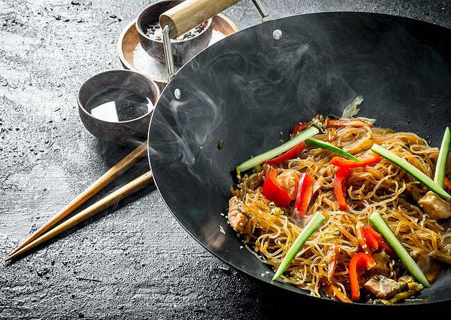 Große Pfanne mit dampfender asiatischer Nudelpfanne, die mit Glasnudeln, dünnen Scheiben von Fleisch, grünen und roten Paprikastreifen sowie Frühlingszwiebeln gefüllt ist.