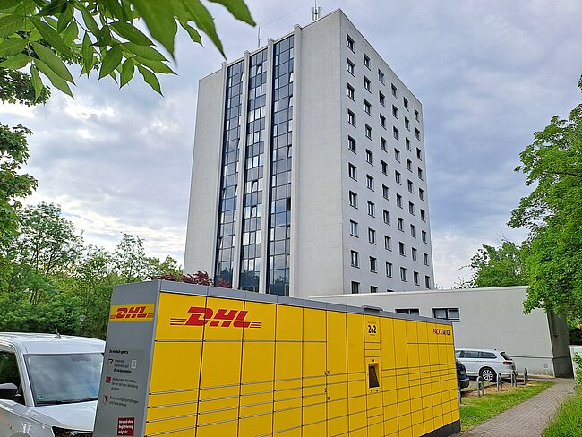 Ein gelber DHL-Lieferwagen steht vor einem mehrstöckigen Gebäude. Im Hintergrund ist ein grauer Wolkenhimmel mit einigen Bäumen zu sehen. Das Gebäude hat viele Fenster und wirkt modern.