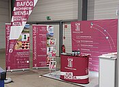 Ein Messe- oder Informationsstand mit Plakaten und Flyern zu Themen wie BAföG, Wohnen, Mensa und Kita. Der Stand ist mit einem roten Tisch und Informationsmaterial ausgestattet. Die Veranstaltung findet am 22. Februar 2024 statt.