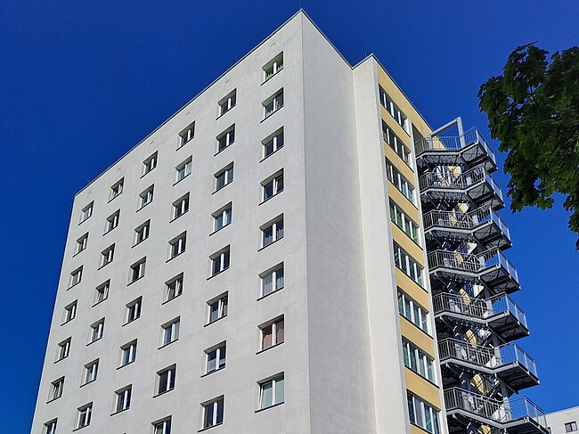 Ein mehrstöckiges Wohngebäude mit vielen Fenstern und einer Außentreppe an der Seite. Das Gebäude hat eine helle Fassade und befindet sich in einer ruhigen Umgebung mit einigen Fahrrädern und Pflanzen im Vordergrund.