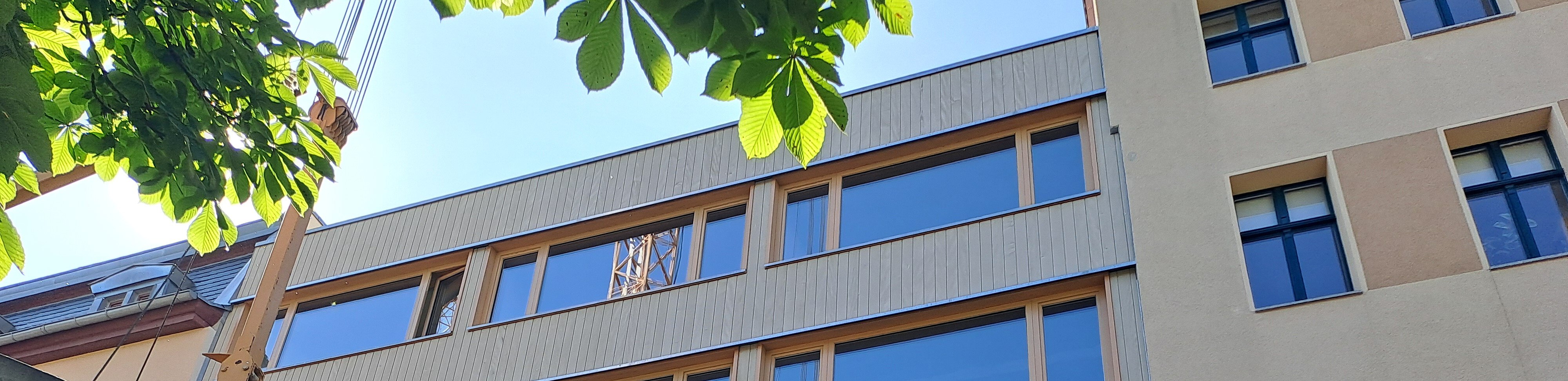 Moderne mehrstöckige Gebäudeecke mit großen, rechteckigen Fenstern und Holzrahmen, helle Fassade mit grauen und beigen Tönen, im Hintergrund Bäume und ein Teil eines weiteren Gebäudes.