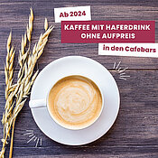 "Ab 2024: Kaffee mit Haferdrink ohne Aufpreis in den Cafebars. Ein Bild zeigt eine Tasse Kaffee mit Hafermilchschaum und Haferhalmen daneben auf einem Holztisch."