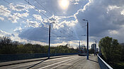 Eine Brücke mit Straßenbahnschienen und Oberleitungen unter einem bewölkten Himmel mit Sonnenlicht, das durch die Wolken scheint. Im Hintergrund sind Bäume und Gebäude zu sehen. Die Brücke ist leer, mit Ausnahme eines fernen Straßenbahnwagens.