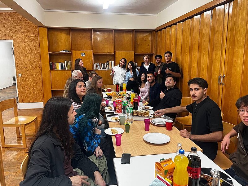 Eine Gruppe von etwa 16 Personen sitzt und steht in einem gemütlichen Raum mit Holzregalen und isst gemeinsam an einem langen Tisch. Es gibt Teller, Getränke und eine lockere, gesellige Atmosphäre.