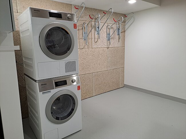 Eine weiße Waschmaschine und ein Wäschetrockner übereinander gestapelt, stehen in einer hellen Waschküche. Im Hintergrund hängen mehrere Hängeparker für Fahrräder an einer Wand.