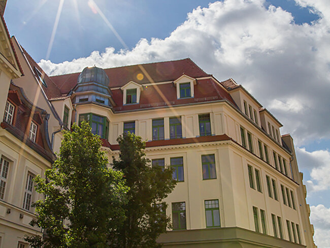 Ein sonniger Tag mit einem historischen Gebäude im Vordergrund. Das Gebäude hat mehrere Stockwerke mit großen Fenstern und einem roten Dach. Vor dem Gebäude stehen Fahrräder und ein Baum. Der Himmel ist blau mit einigen weißen Wolken.