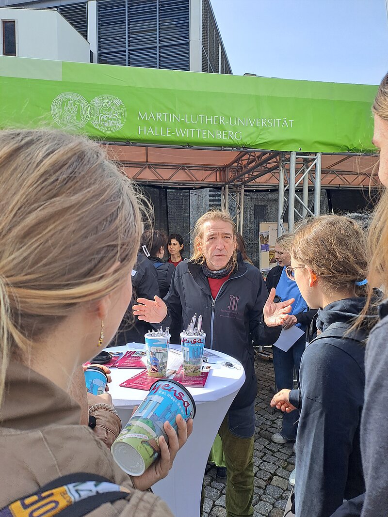 Eine Gruppe von Menschen steht im Freien und unterhält sich. Im Hintergrund ist ein Banner der Martin-Luther-Universität Halle-Wittenberg zu sehen. Eine Frau spricht mit anderen Personen, während eine weitere Person ein Getränk in der Hand hält. Die Atmosphäre wirkt informell und gesellig.