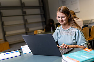 Eine Frau mit Brille sitzt an einem Tisch und arbeitet konzentriert an einem Laptop. Vor ihr liegen Bücher und Notizen. Sie trägt ein helles Oberteil und lächelt leicht. Der Raum wirkt wie ein ruhiger Arbeits- oder Studienbereich.