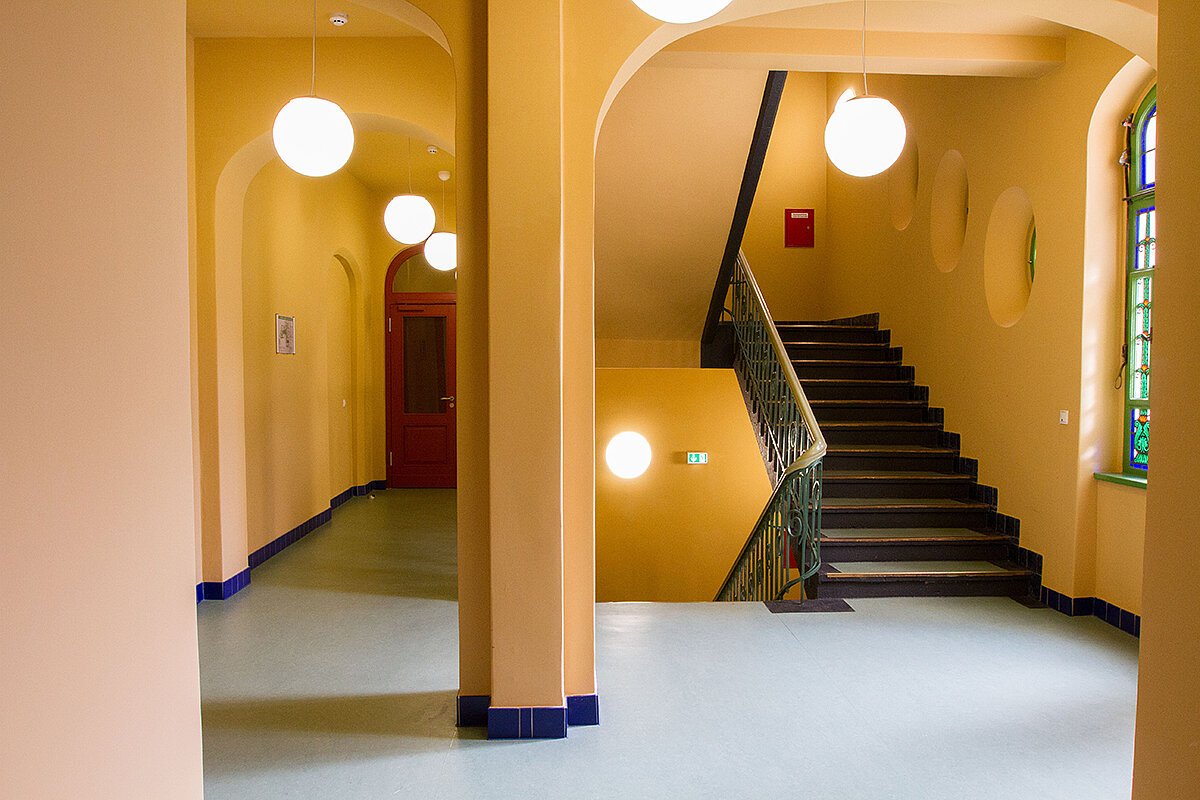 Ein heller Flur mit gelben Wänden und mehreren runden Deckenlampen. Links befindet sich ein langer Korridor mit einer roten Tür am Ende. Rechts führt eine Treppe mit einem gelben Handlauf nach oben. Große Fenster lassen Tageslicht herein.