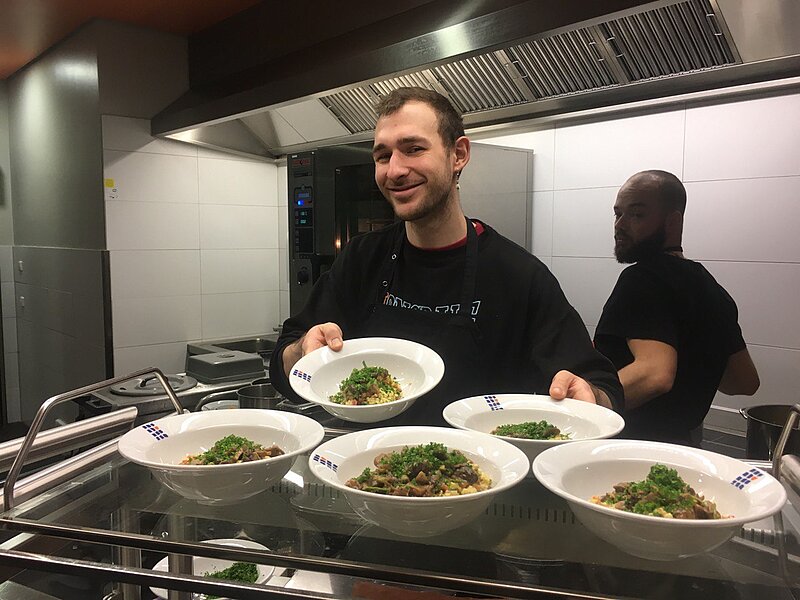 Zwei Köche in einer Küche bereiten mehrere Teller mit Essen vor. Der vordere Koch lächelt und hält einen Teller mit einem grünen Topping. Die Küche ist modern mit Edelstahlgeräten und einer Dunstabzugshaube.