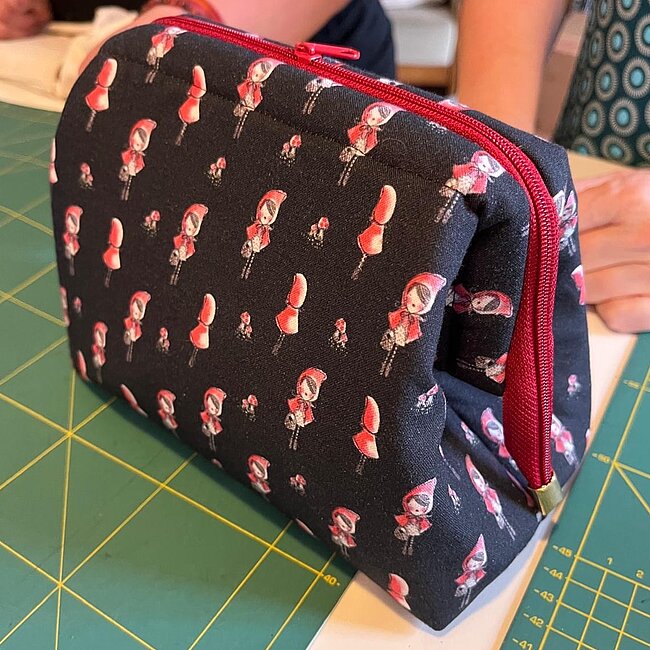 Kulturtasche, Foto: Cordelia Minuth, Nähzimmer Schwarze Reisetasche mit rot-pinkem Muster aus fliegenden Flamingos, offen liegend auf grüner Schnittunterlage, mit rotem Reißverschluss.