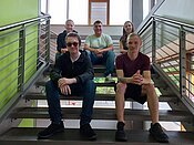 Eine Gruppe von fünf Personen sitzt auf einer Treppe in einem modernen Gebäude. Zwei Männer sitzen vorne auf den Stufen, während drei weitere Personen hinter ihnen auf einer höheren Stufe sitzen. Alle lächeln und wirken entspannt.