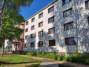 Ein mehrstöckiges Wohngebäude mit weißen Wänden und vielen rechteckigen Fenstern. Vor dem Gebäude befindet sich ein gepflegter Garten mit Bäumen und Sträuchern. Der Himmel ist klar und blau.