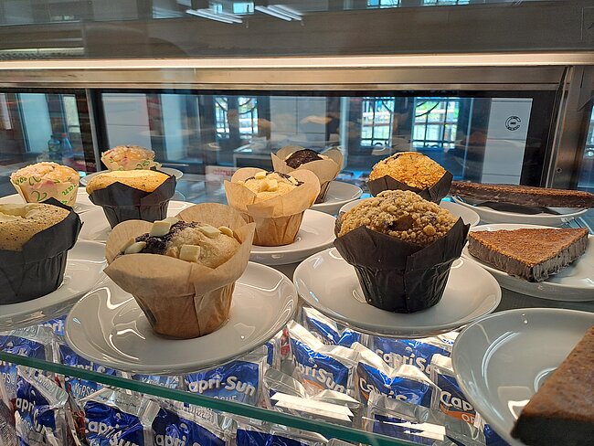 Ausstellung von sechs verschiedenen Muffins in Glastheke: zwei mit Nussstreusel, einer mit Schokostückchen, einer mit Aprikosenglasur, einer mit karamellisierter Oberfläche und zwei klassisch braun mit unterschiedlichen Texturen. Dazu ein Stück Kuchen auf weißer Platte.