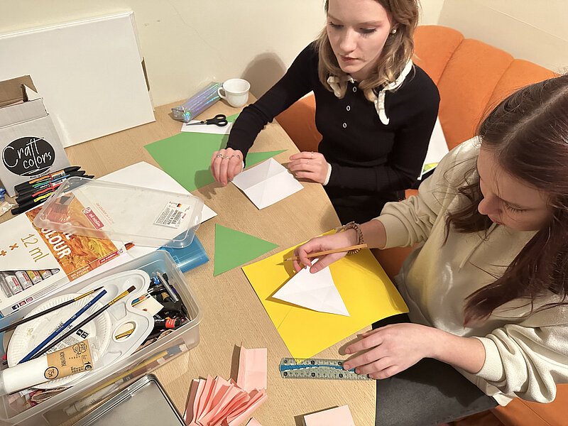 Zwei Frauen sitzen an einem Tisch und basteln mit Papier und Scheren. Verschiedene Bastelmaterialien wie bunte Papiere, Stifte und Kleber sind auf dem Tisch verteilt. Sie scheinen konzentriert bei der Arbeit zu sein.