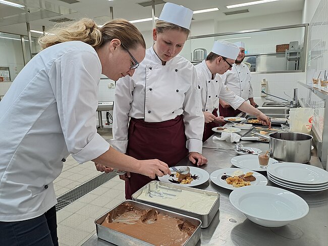 Koch-Ausbilderin und Koch-Auszubildende in einer professionellen Küche: Zwei Personen in Kochjacken und eine Person in Kochuniform mit Hut dekorieren gemeinsam einen Dessertteller, während im Hintergrund weitere Köche arbeiten und Geschirr bereitsteht.