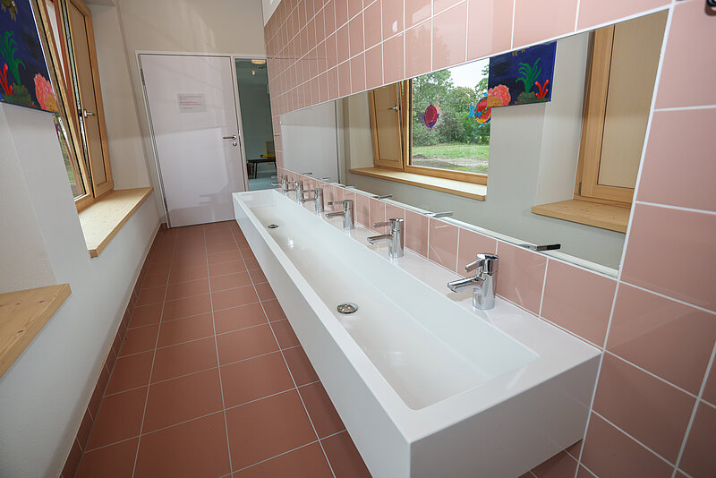 Ein sauberes, modernes Badezimmer mit mehreren Waschbecken und Spiegeln. Die Wände sind mit rosa Fliesen verkleidet, und es gibt ein Fenster mit Blick auf Blumen. Der Boden ist mit rosa Fliesen belegt. Es gibt auch einen weißen Schrank und eine Tür.