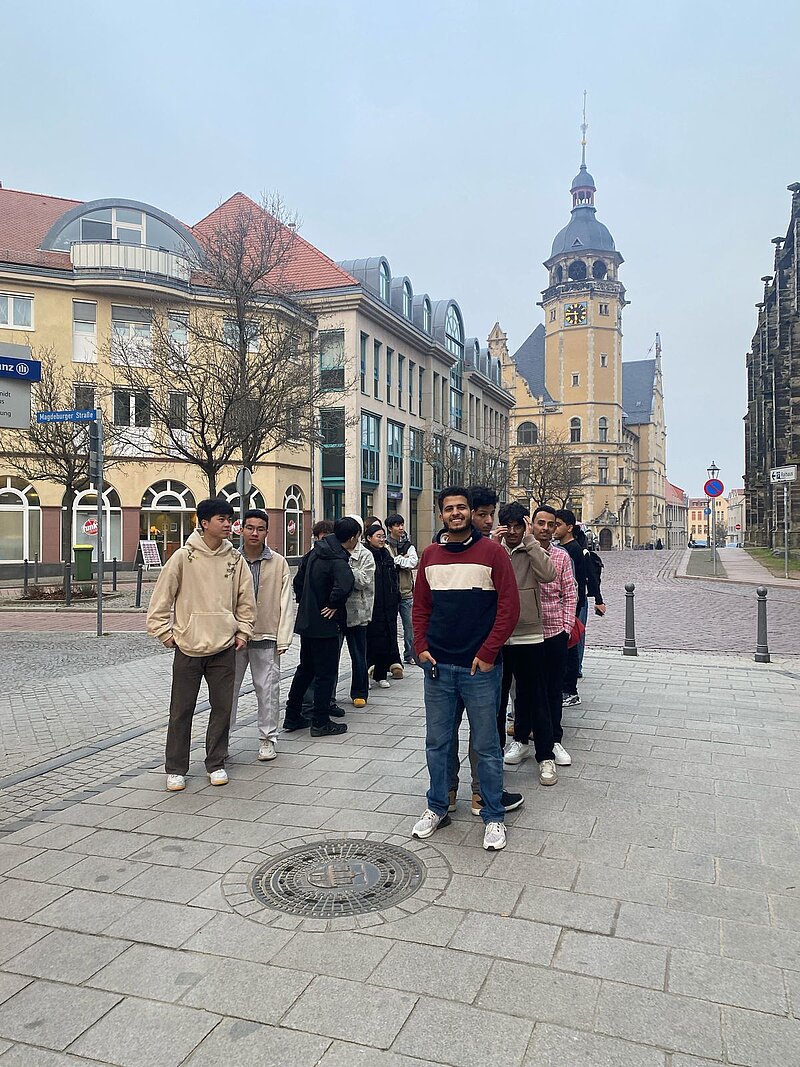 Eine Gruppe von Menschen steht auf einem gepflasterten Platz vor einem historischen Gebäude mit einem Turm. Im Hintergrund sind weitere Gebäude und Bäume zu sehen. Die Personen scheinen sich zu unterhalten und die Umgebung zu genießen.