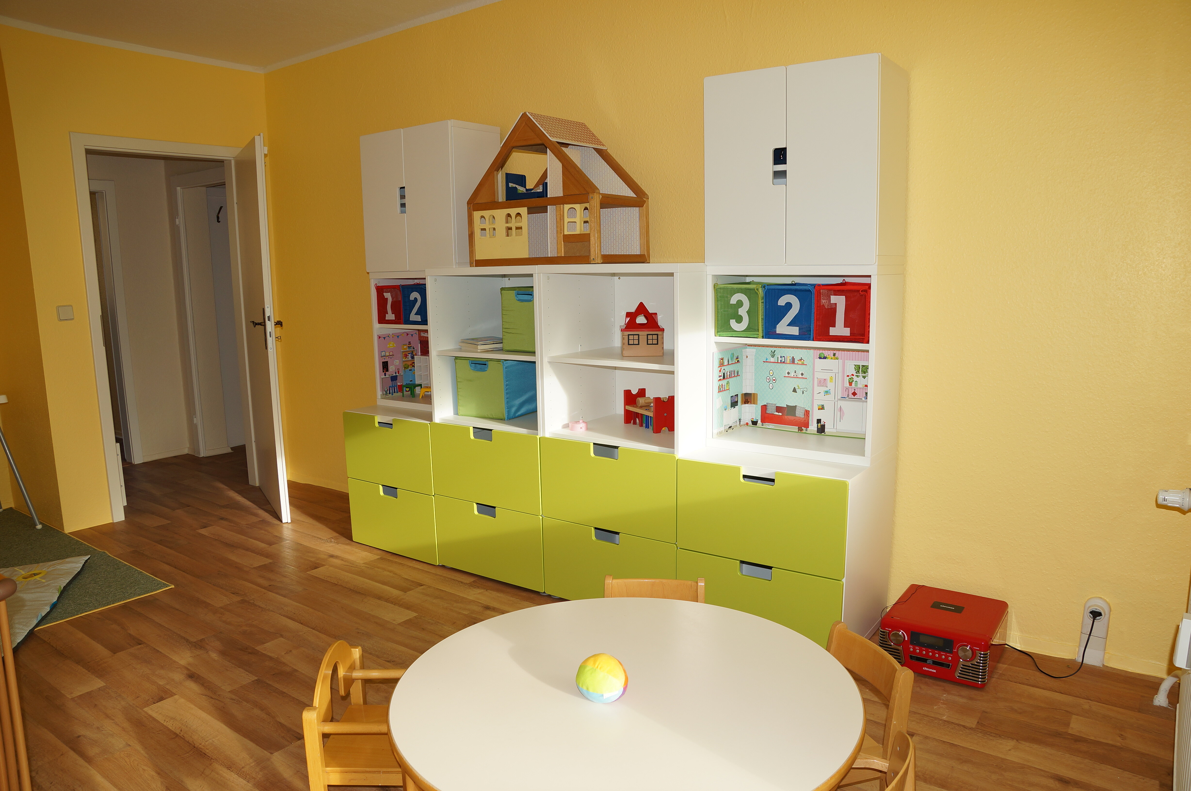 Ein Kinderzimmer mit einem weißen Tisch und kleinen Stühlen. An der Wand steht ein weißes und grünes Regal mit Spielzeug, Büchern und einer kleinen Holzhütte. Der Boden ist aus Holz und die Wände sind gelb gestrichen.