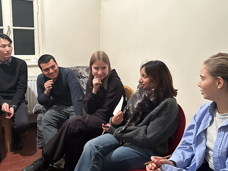 Eine Gruppe von fünf Personen sitzt in einem Raum und unterhält sich. Sie wirken entspannt und konzentriert. Die Personen sind unterschiedlich gekleidet und sitzen auf Stühlen und einer Couch. Der Raum hat helle Wände und ein Fenster.